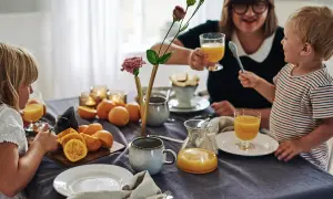 Familjen samlas kring frukostbordet med apelsinjuice och frukt. Duken är av linne, redo att stryka för nästa måltid.