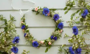 En krans av midsommar blommor med blåklint och rosa rosor, omgiven av vilda blommor på en ljusblå träbakgrund.