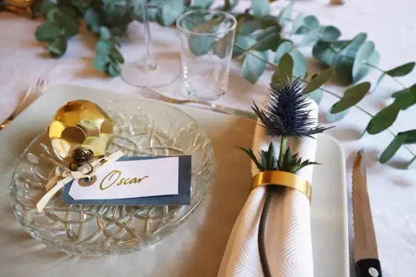 Elegant nyårsdukning med gulddekorationer och en namnskylt för Oscar. En gyllene kotte pryder tallriken, medan en blå tistel med guldfärgad servettring skapar en vacker detalj.